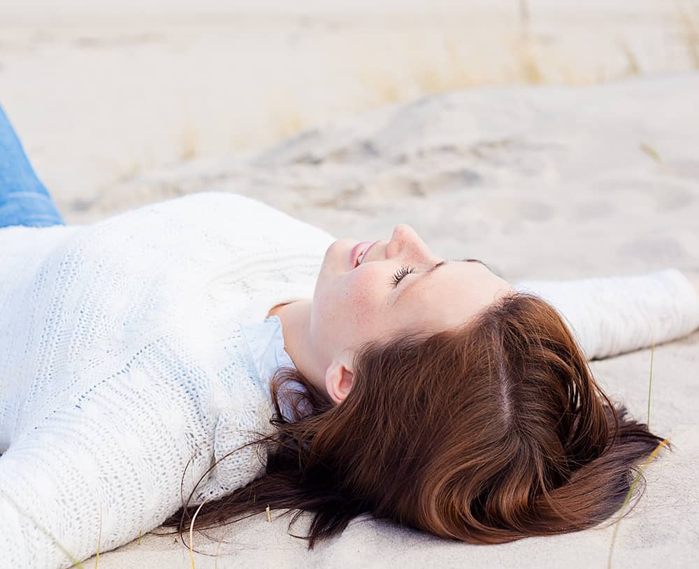 Eine mit ausgebreiteten Armen im Sand liegende junge Frau, die in den Himmel schaut.